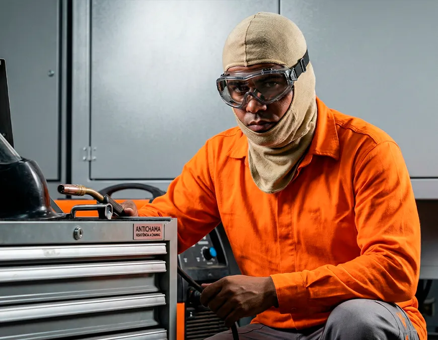 Soldador industrial vestindo camisa laranja, &oacute;culos de seguran&ccedil;a e balaclava bege antichama, segurando tocha de solda pr&oacute;ximo a um carrinho de ferramentas.