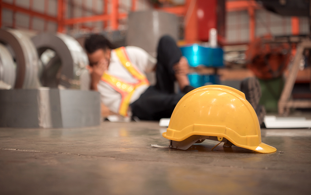 Capacete de seguran&ccedil;a amarelo ca&iacute;do em primeiro plano no ch&atilde;o de uma f&aacute;brica, com um trabalhador de colete reflexivo ao fundo desfocado, sentado e segurando o joelho em sinal de dor ap&oacute;s um acidente.