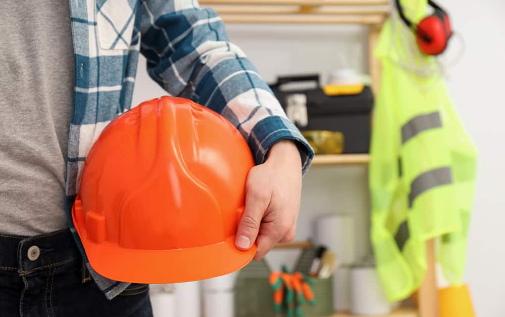 pessoa segurando um capacete de seguran&ccedil;a laranja, com equipamentos e um colete refletivo desfocados ao fundo.