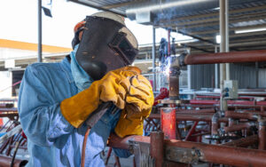 Trabalhador industrial de macacão alaranjado e óculos transparentes ajustando a jugular de um capacete de proteção amarelo na cabeça, em um galpão com outros capacetes pendurados na parede.