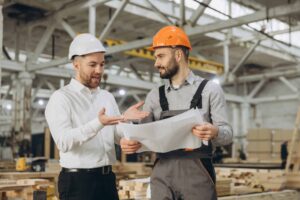 Dois engenheiros examinam plantas em uma fábrica de construções modulares, discutindo planos de construção e detalhes do projeto.