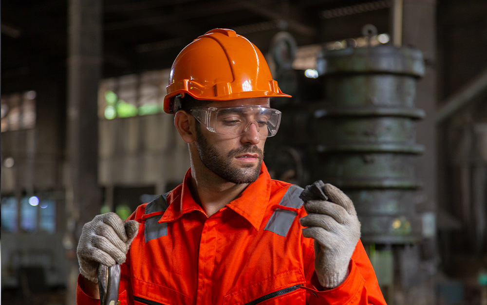 Trabalhador industrial de capacete laranja, óculos de segurança e macacão alaranjado, usando luvas e examinando com atenção um pequeno pedaço de material escuro nas mãos.