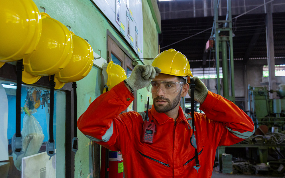 Trabalhador industrial de capacete laranja, óculos de segurança e macacão alaranjado, usando luvas e examinando com atenção um pequeno pedaço de material escuro nas mãos.
