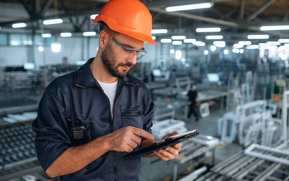 Trabalhador de uniforme escuro, capacete laranja e &oacute;culos de seguran&ccedil;a manuseando um tablet dentro de uma f&aacute;brica