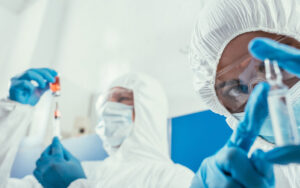 Vista de ângulo baixo de dois cientistas em trajes completos de proteção (macacão branco, máscaras faciais e óculos de segurança). O cientista em primeiro plano está segurando uma pequena ampola de vidro transparente com luvas azuis, com o rosto e os olhos nítidos olhando para a câmera. O segundo cientista está desfocado ao fundo à esquerda, usando uma seringa para retirar líquido de um frasco pequeno contendo uma solução laranja. O fundo do laboratório é composto por superfícies clínicas e um painel azul desfocado.