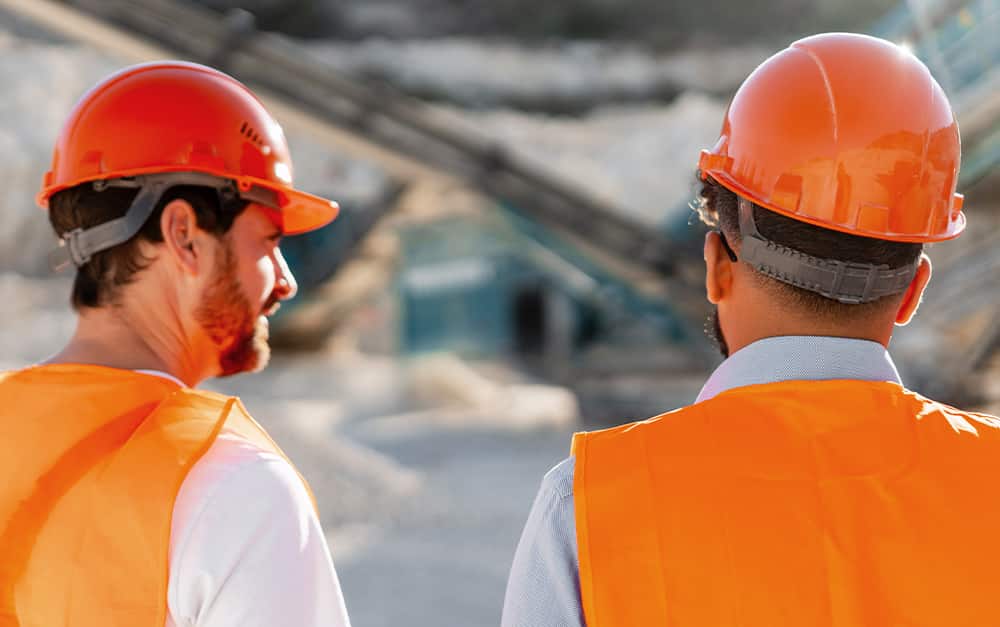 Dois trabalhadores da constru&ccedil;&atilde;o civil de capacetes e coletes laranjas vistos de costas