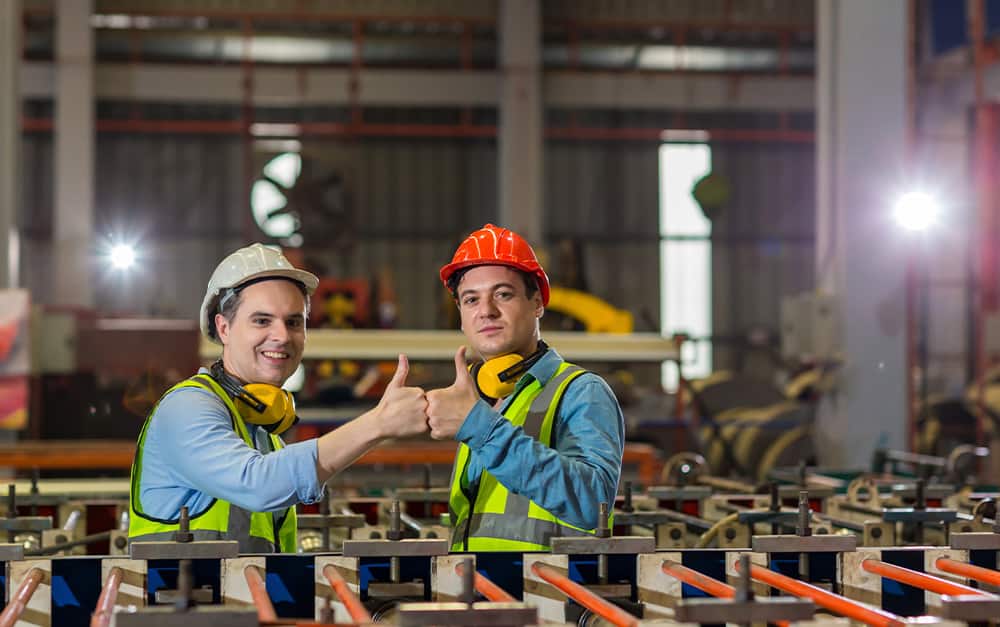 Dois trabalhadores de f&aacute;brica de capacetes brancos e coletes verdes d&atilde;o um duplo joinha em frente a m&aacute;quinas industriais.