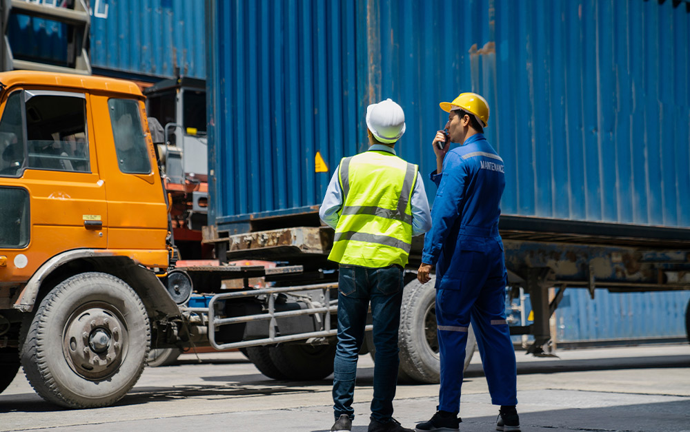 Dois trabalhadores de porto de cont&ecirc;ineres, um em capacete branco e colete de seguran&ccedil;a e outro em capacete amarelo e macac&atilde;o azul, conversam por r&aacute;dio perto de um caminh&atilde;o laranja e cont&ecirc;ineres de carga azuis.
