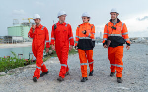 Quatro profissionais de segurança e engenharia vestindo uniformes laranja de alta visibilidade com faixas reflexivas e capacetes brancos, caminhando lado a lado em uma área industrial externa.
