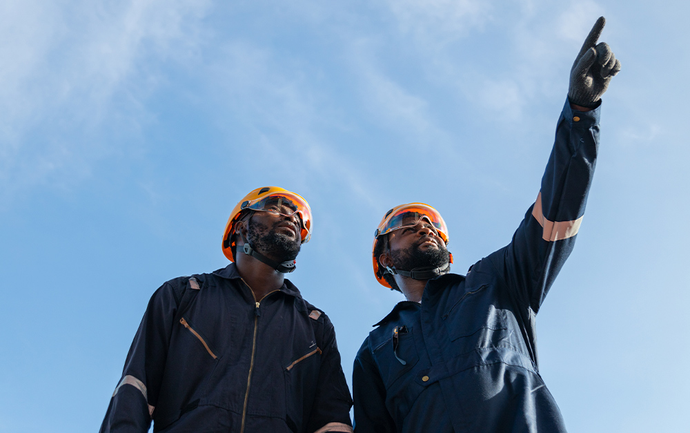 Dois trabalhadores em foto tirada de baixo para cima, vestindo macac&otilde;es azul-escuro, capacetes amarelos e &oacute;culos de prote&ccedil;&atilde;o, com um deles apontando para cima contra um c&eacute;u azul limpo.