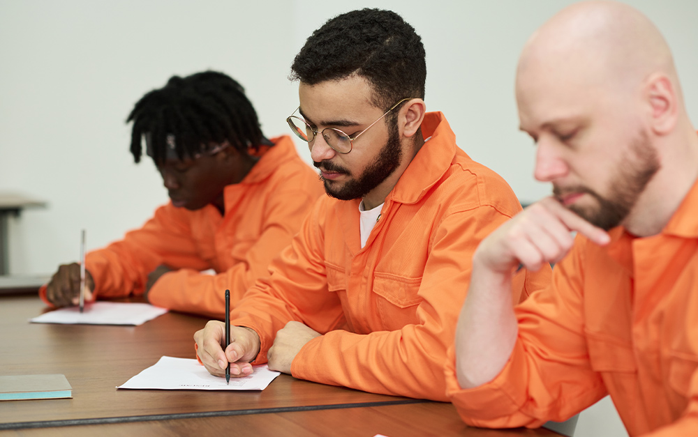 Tr&ecirc;s homens vestindo macac&otilde;es alaranjados de trabalho sentados a uma mesa de madeira, concentrados enquanto realizam uma prova ou preenchem formul&aacute;rios.