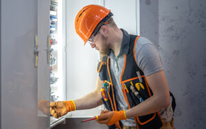 Eletricista com capacete laranja, óculos de segurança e colete de ferramentas realizando manutenção em um painel elétrico aberto com disjuntores.