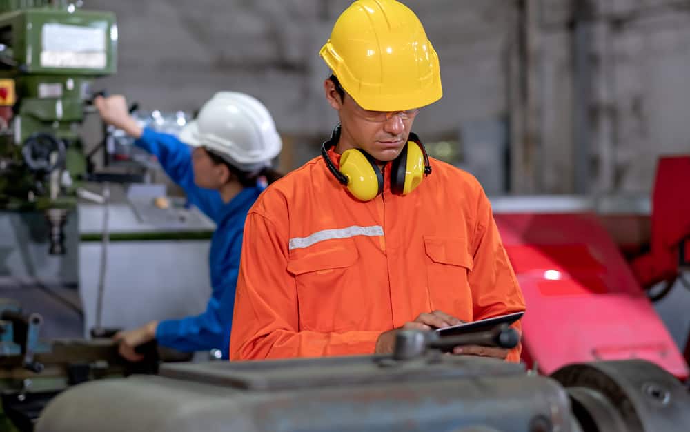 Trabalhador industrial de macac&atilde;o laranja, capacete amarelo e protetores auriculares consultando um tablet, com outra funcion&aacute;ria operando uma m&aacute;quina ao fundo.