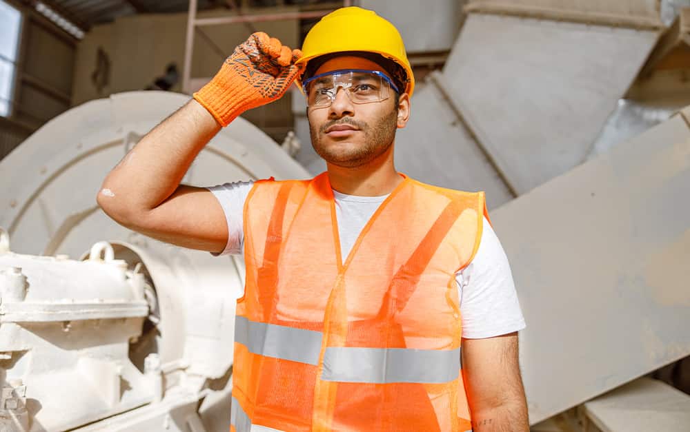 Trabalhador usando capacete amarelo, &oacute;culos de seguran&ccedil;a, colete refletivo laranja e luvas, com uma das m&atilde;os apoiada na aba do capacete em um ambiente industrial.
