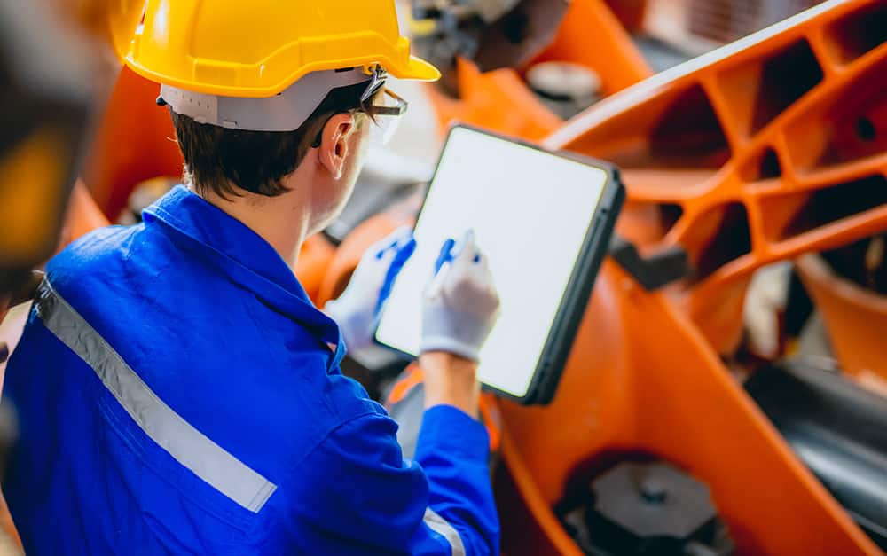 um trabalhador de uniforme azul e capacete amarelo usando uma caneta touch na tela em branco de um tablet, pr&oacute;ximo a uma estrutura met&aacute;lica