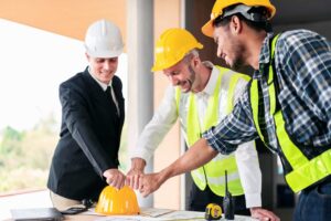 Três homens estão em pé ao redor de uma mesa, um deles vestindo um colete amarelo
